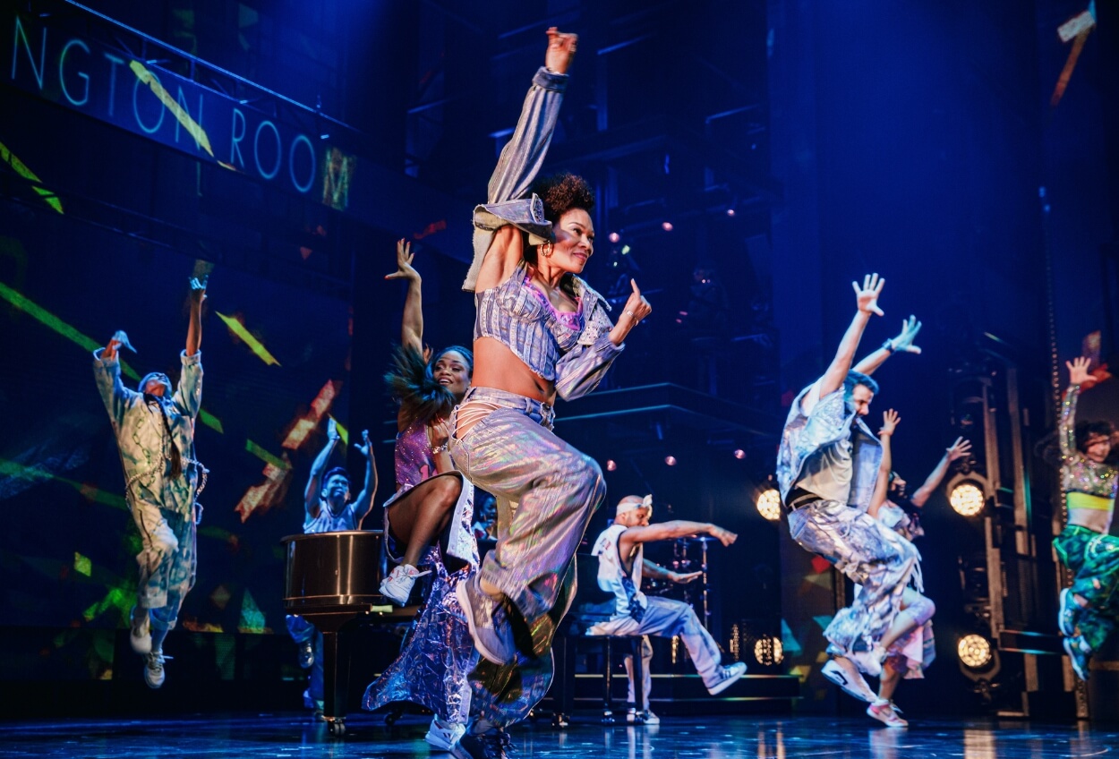A dynamic scene from a theatrical production featuring a group of performers energetically dancing on stage with colorful lighting and a vibrant backdrop.